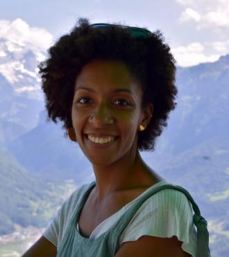 Femme souriante aux cheveux afro et boucles d'oreilles dorées, posant devant des montagnes.