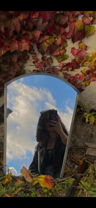 Autoportrait en miroir d'une personne aux cheveux bouclés se cachant le visage derrière un téléphone, le miroir reflétant un ciel bleu nuageux et étant entouré de feuilles rouges et jaunes.